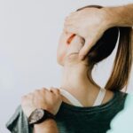 Back view of crop faceless chiropractor in wristwatch stretching neck of unrecognizable female patient during medical examination in doctor office in clinic